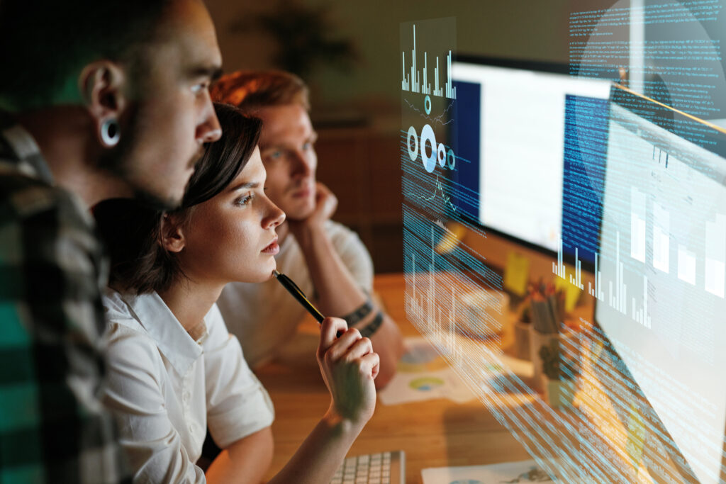 Three people working on a computer screen, a young woman in the middle has a pen in her hand, which is touching her chin, she looks to be in thought.